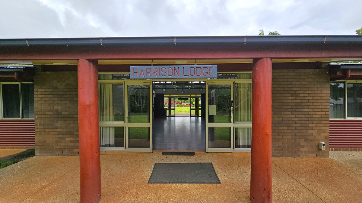 A road with white markings on the left, flanked by trees and bollards. On the right, a low-rise building with a brown brick facade and pitched, extended roofing. Well-manicured green grass lines its left side. Trees and shrubs surround the area. A road with white markings on the left, flanked by trees and bollards. On the right, a low-rise building with a brown brick facade and pitched, extended roofing. Well-manicured green grass lines its left side. Trees and shrubs surround the area.