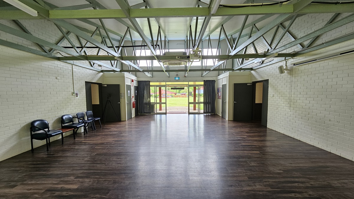 A spacious open area with a brown timber floor. Black plastic chairs arranged against the left wall. White brick wall and white ceiling with exposed metal beams. Black outward-opening doors on both sides lead to corridors. Further back, green grass is visible. A spacious open area with a brown timber floor. Black plastic chairs arranged against the left wall. White brick wall and white ceiling with exposed metal beams. Black outward-opening doors on both sides lead to corridors. Further back, green grass is visible.