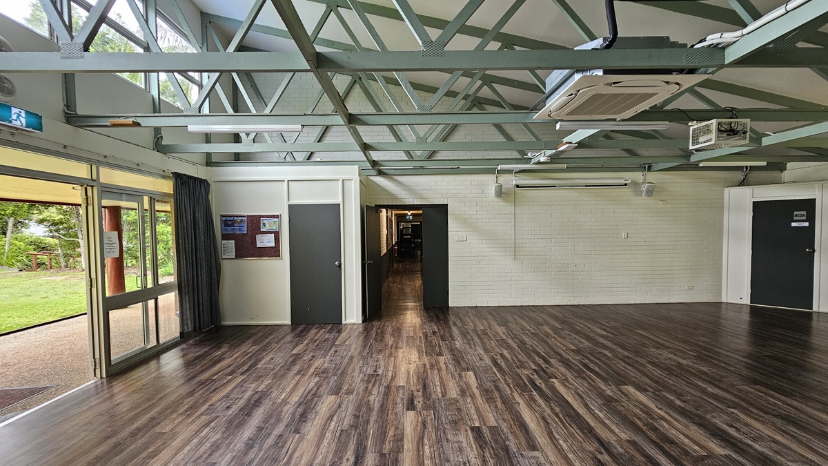 A spacious open area with a brown timber floor. On the left. a white brick wall holds a brown board with posters. The white ceiling features exposed metal beams, fluorescent lights, and air conditioning. A black door on the left leads to a corridor, and another black door is on the right. To the left, the open glass door exposes green grass outside. A spacious open area with a brown timber floor. On the left. a white brick wall holds a brown board with posters. The white ceiling features exposed metal beams, fluorescent lights, and air conditioning. A black door on the left leads to a corridor, and another black door is on the right. To the left, the open glass door exposes green grass outside.