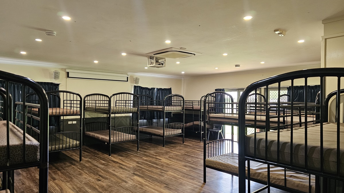 A room with a brown timber floor featuring metal bunk beds strategically positioned against the walls, each equipped with mattresses. The white ceiling has LED lights, air conditioning, and a projector machine. The wall is white with a projector board. Windows are covered with dark blue curtains. A room with a brown timber floor featuring metal bunk beds strategically positioned against the walls, each equipped with mattresses. The white ceiling has LED lights, air conditioning, and a projector machine. The wall is white with a projector board. Windows are covered with dark blue curtains.