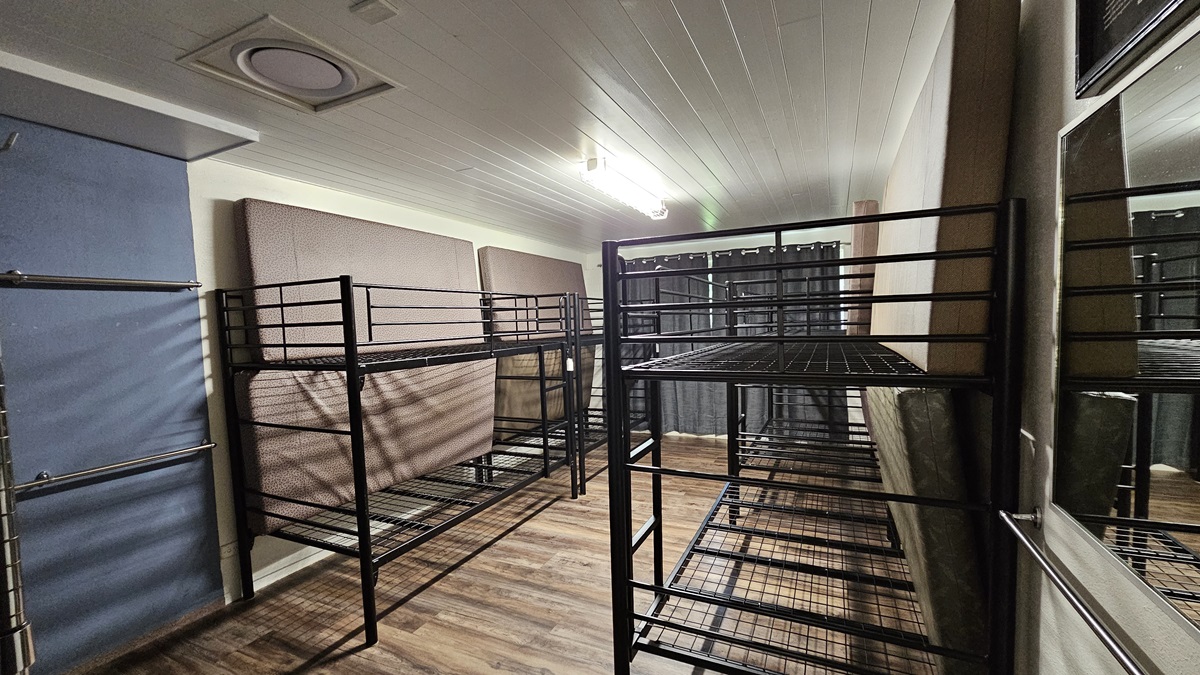 A room with a brown timber floor featuring black metal bunk beds strategically positioned against the walls, each equipped with mattresses laid on the sides. The white ceiling has LED lights. The wall is all white. A room with a brown timber floor featuring black metal bunk beds strategically positioned against the walls, each equipped with mattresses laid on the sides. The white ceiling has LED lights. The wall is all white.