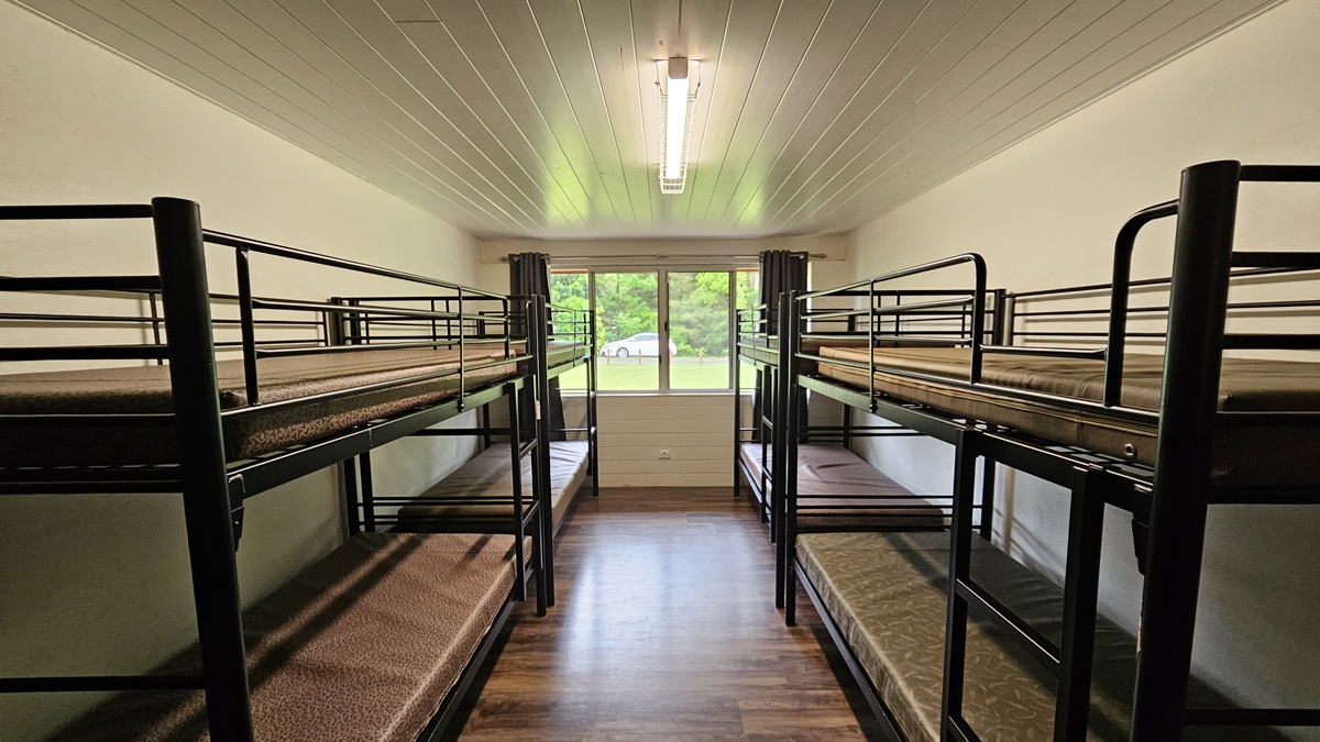 A room with a brown timber floor featuring black metal bunk beds strategically positioned against the walls, each equipped with mattresses. The white ceiling has a fluorescent light. In the middle is a glass window with dark curtains rolled to the sides, offering a view of the green grass outside and a white car. A room with a brown timber floor featuring black metal bunk beds strategically positioned against the walls, each equipped with mattresses. The white ceiling has a fluorescent light. In the middle is a glass window with dark curtains rolled to the sides, offering a view of the green grass outside and a white car.