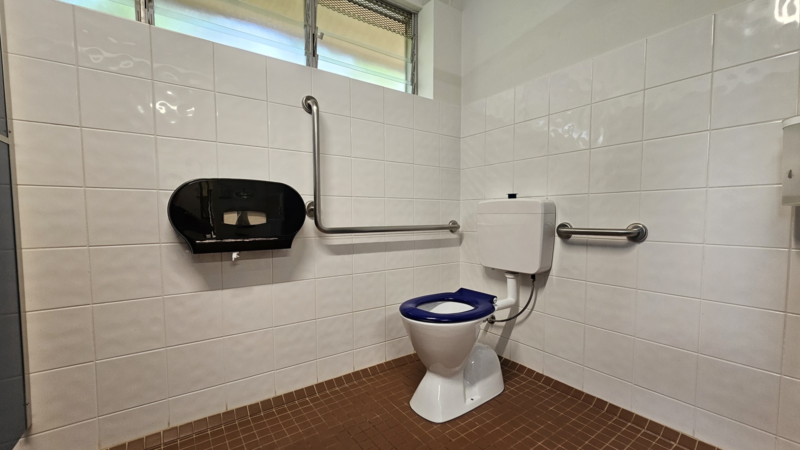 In the corner, a toilet bowl with handrails. A tissue dispenser is on the left wall. Glass jalousie windows are on the upper left, and a liquid soap dispenser is on the upper right wall. White tiles cover the walls, while the floor is tiled brown. In the corner, a toilet bowl with handrails. A tissue dispenser is on the left wall. Glass jalousie windows are on the upper left, and a liquid soap dispenser is on the upper right wall. White tiles cover the walls, while the floor is tiled brown.