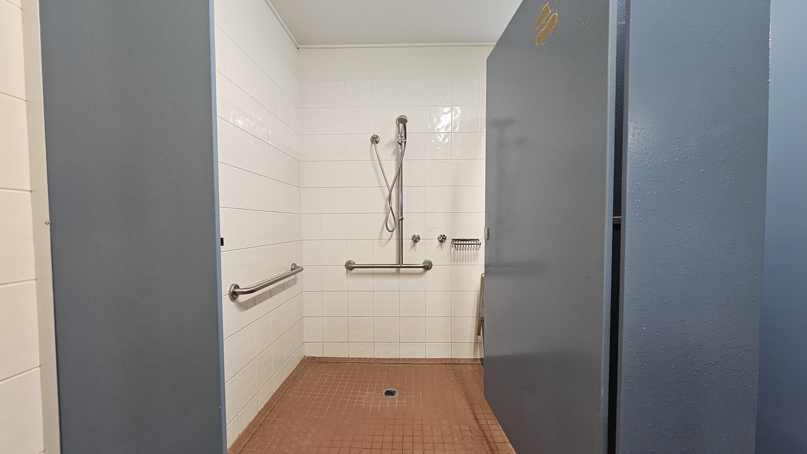 A shower with hand grabs and a soap holder mounted on the wall. White tiles cover the wall, while brown tiles adorn the floor. The ceiling is white, and the door is grey. A shower with hand grabs and a soap holder mounted on the wall. White tiles cover the wall, while brown tiles adorn the floor. The ceiling is white, and the door is grey.