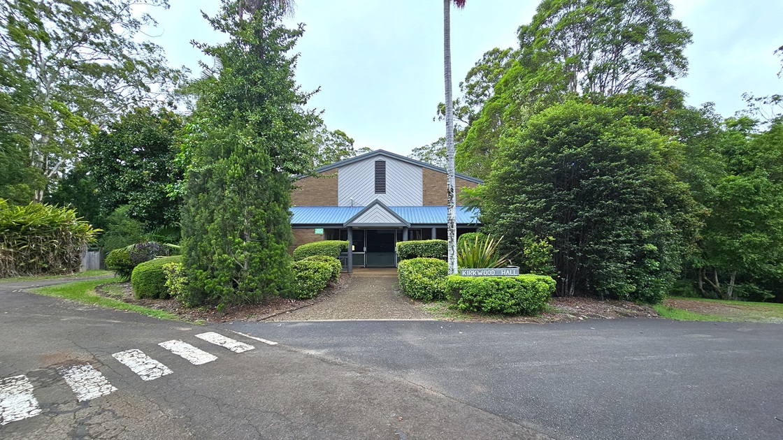 In the middle is a low-rise facility with a pitched roof, and a brown brick facade with white and blue trims. Trimmed shrubs border the front and sides. A sign is placed next to a tall wooden post on the right. Various trees surround the area and border the road with white markings for pedestrians. In the middle is a low-rise facility with a pitched roof, and a brown brick facade with white and blue trims. Trimmed shrubs border the front and sides. A sign is placed next to a tall wooden post on the right. Various trees surround the area and border the road with white markings for pedestrians.