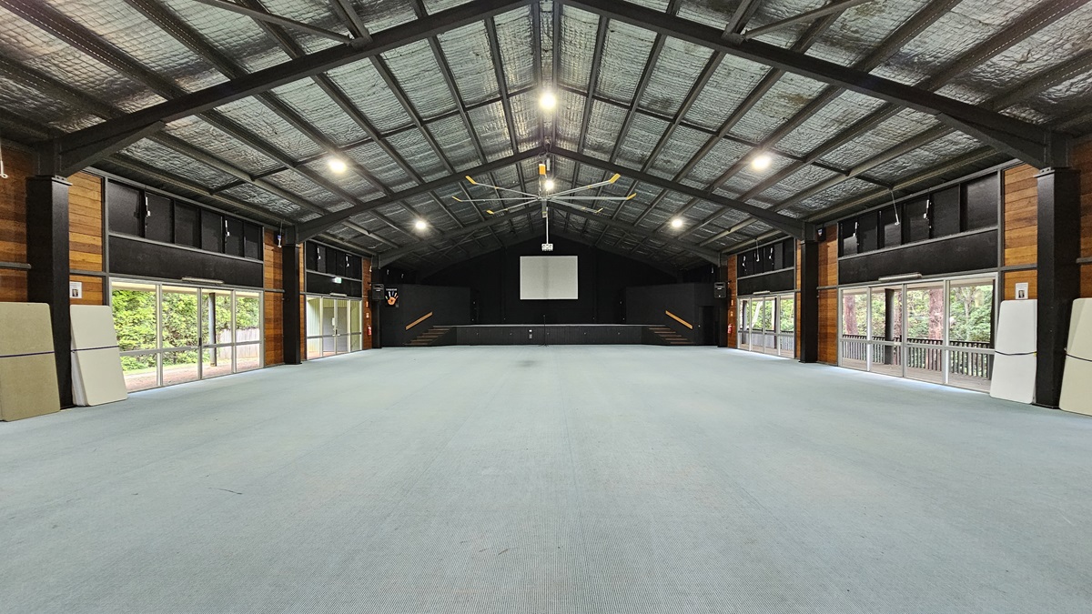 A spacious open area with a raised platform in the middle and a projector screen. The ceiling is pitched with an industrial look, featuring exposed metal beams, silver insulation, and a large ceiling fan. Both sides have large glass windows and doors offering a view of the trees outside. The wall features black and brown timber. The floor is covered with grey tiles. A spacious open area with a raised platform in the middle and a projector screen. The ceiling is pitched with an industrial look, featuring exposed metal beams, silver insulation, and a large ceiling fan. Both sides have large glass windows and doors offering a view of the trees outside. The wall features black and brown timber. The floor is covered with grey tiles.