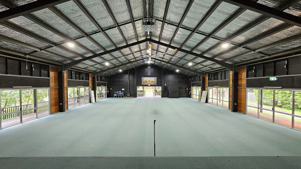 A spacious open area with a pitched industrial-style ceiling, exposed metal beams, silver insulation, a large ceiling fan, and a projector machine. Both sides have large glass windows and doors offering a view of trees outside. The walls feature black and brown timber, and the floor is covered with grey tiles. Plastic chairs are piled in the left corner, and a large flatscreen TV is mounted on the middle wall parallel to the entrance. A spacious open area with a pitched industrial-style ceiling, exposed metal beams, silver insulation, a large ceiling fan, and a projector machine. Both sides have large glass windows and doors offering a view of trees outside. The walls feature black and brown timber, and the floor is covered with grey tiles. Plastic chairs are piled in the left corner, and a large flatscreen TV is mounted on the middle wall parallel to the entrance.