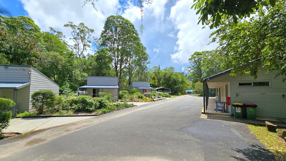In the middle is a concrete road. Low-rise buildings with concrete walls and corrugated roofing are positioned on both sides. The longer building on the right displays a fire extinguisher on the wall, along with a table, chair, and two rubbish bins. Trees and grass surround the area. In the middle is a concrete road. Low-rise buildings with concrete walls and corrugated roofing are positioned on both sides. The longer building on the right displays a fire extinguisher on the wall, along with a table, chair, and two rubbish bins. Trees and grass surround the area.