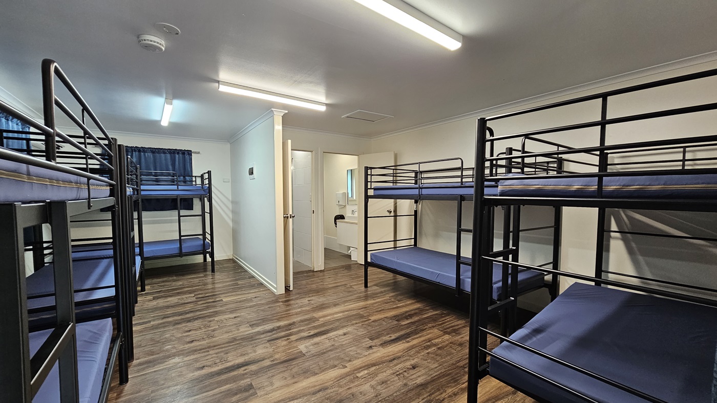A room with metal bunk beds, each with mattresses. Large windows with grey curtains rolled to the sides. A mirror and signage are mounted on the middle wall. White walls and ceiling with fluorescent lights and air conditioning. Brown timber floor. A room with metal bunk beds, each with mattresses. Large windows with grey curtains rolled to the sides. A mirror and signage are mounted on the middle wall. White walls and ceiling with fluorescent lights and air conditioning. Brown timber floor.