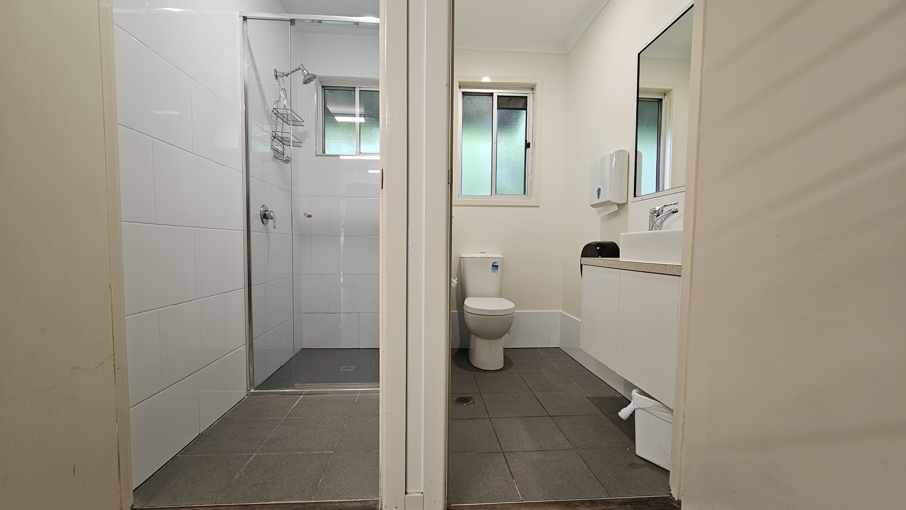 Two separate cubicles. To the right, a toilet bowl is positioned in the corner with a glass window above. On the right wall, a mirror, tissue dispenser, and hand basin are mounted. A rubbish bin is placed underneath. To the left is a shower area enclosed with glass. Both cubicles feature white walls, grey floors, and small windows. Two separate cubicles. To the right, a toilet bowl is positioned in the corner with a glass window above. On the right wall, a mirror, tissue dispenser, and hand basin are mounted. A rubbish bin is placed underneath. To the left is a shower area enclosed with glass. Both cubicles feature white walls, grey floors, and small windows.