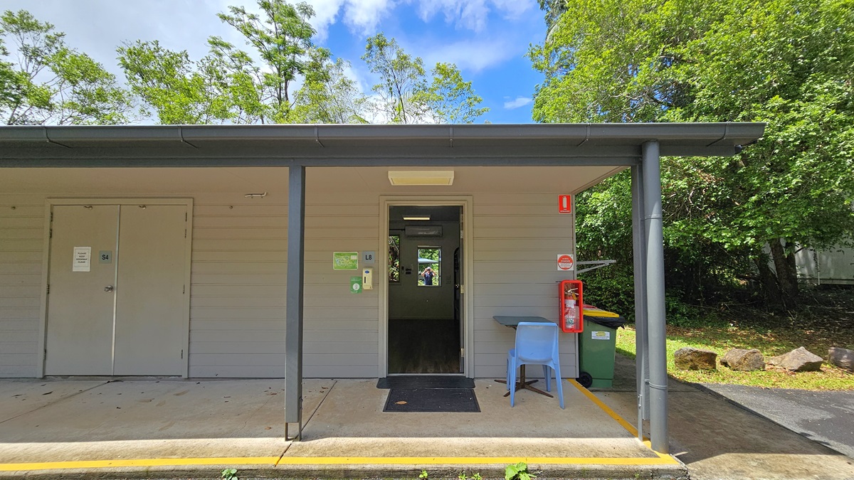 A low-rise building with extended roofing and white brick walls with multiple doors. The open door on the right reveals timber floors and a mirror inside. The outer wall features a fire extinguisher and signage. A table and plastic chair are placed to the right of the door. Trees and grass surround the area, and rubbish bins visible on the right. A low-rise building with extended roofing and white brick walls with multiple doors. The open door on the right reveals timber floors and a mirror inside. The outer wall features a fire extinguisher and signage. A table and plastic chair are placed to the right of the door. Trees and grass surround the area, and rubbish bins visible on the right.