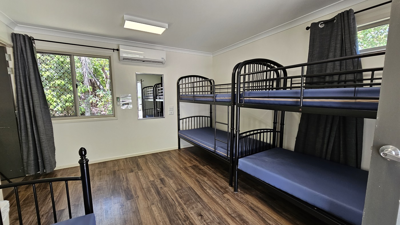 A room with metal bunk beds, each with mattresses. Large windows with grey curtains rolled to the sides. A mirror and signage are mounted on the middle wall. White walls and ceiling with fluorescent lights and air conditioning. Brown timber floor. A room with metal bunk beds, each with mattresses. Large windows with grey curtains rolled to the sides. A mirror and signage are mounted on the middle wall. White walls and ceiling with fluorescent lights and air conditioning. Brown timber floor.