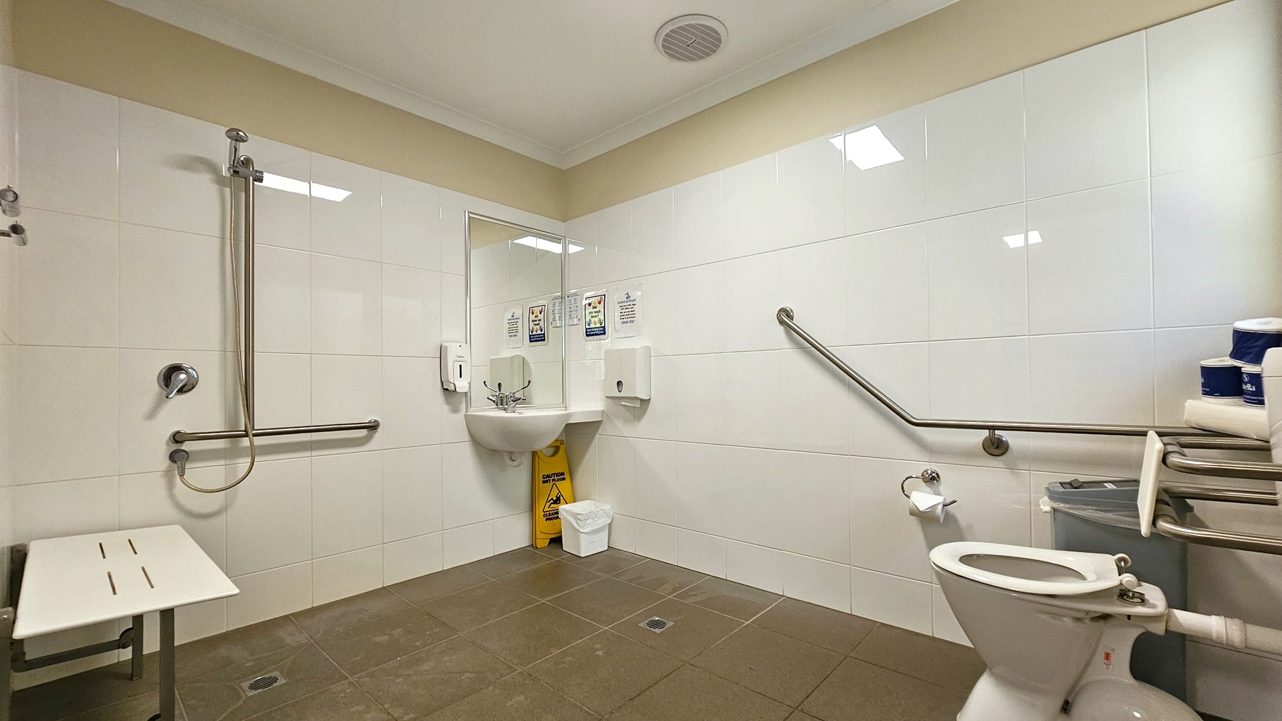 A toilet bowl equipped with handrails on the right. Tissue rolls and a rubbish bin are positioned to the right. In the middle corner is a hand basin. There is a tissue dispenser, liquid soap dispenser, and stickers. A rubbish bin and a yellow warning sign are underneath. Adjacent is a shower with handrails, and next to it is a foldable bench chair mounted to the left wall. The ceiling is white with a cream accent, and the walls are tiled with white tiles, while the floor has brown tiles. A toilet bowl equipped with handrails on the right. Tissue rolls and a rubbish bin are positioned to the right. In the middle corner is a hand basin. There is a tissue dispenser, liquid soap dispenser, and stickers. A rubbish bin and a yellow warning sign are underneath. Adjacent is a shower with handrails, and next to it is a foldable bench chair mounted to the left wall. The ceiling is white with a cream accent, and the walls are tiled with white tiles, while the floor has brown tiles.