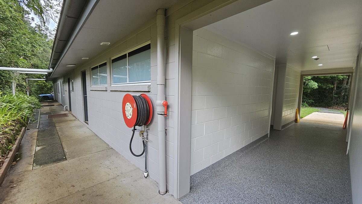 A low-rise building with a white brick facade, multiple doors, and glass windows. A fire hose reel is mounted near the corner. Landscaped ornamental plants and trees border the left side. The lobby on the right has grey tiles and leads to the other side of the building. Safety warning signs are placed near the wall. A low-rise building with a white brick facade, multiple doors, and glass windows. A fire hose reel is mounted near the corner. Landscaped ornamental plants and trees border the left side. The lobby on the right has grey tiles and leads to the other side of the building. Safety warning signs are placed near the wall.