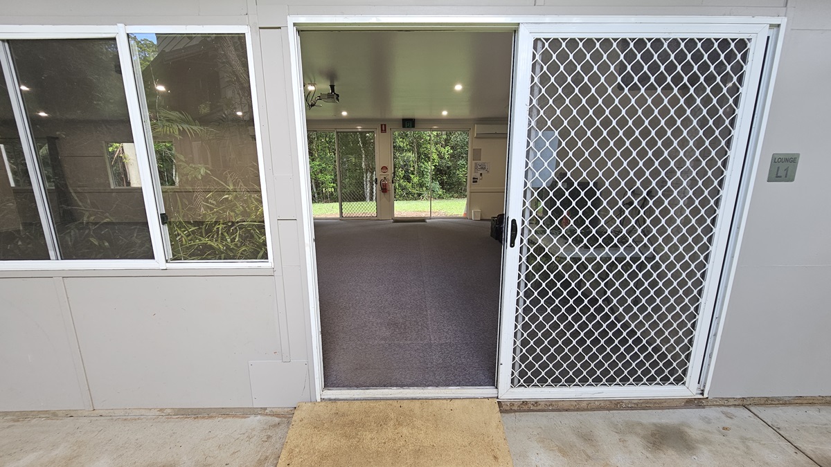 A glass door with a security screen and a yellow mat. Adjacent are glass windows. Inside is a spacious open space with grey flooring and a white ceiling. Signs, along with a fire extinguisher, are mounted on the middle wall, next to another glass door that leads to the green field outside. A glass door with a security screen and a yellow mat. Adjacent are glass windows. Inside is a spacious open space with grey flooring and a white ceiling. Signs, along with a fire extinguisher, are mounted on the middle wall, next to another glass door that leads to the green field outside.