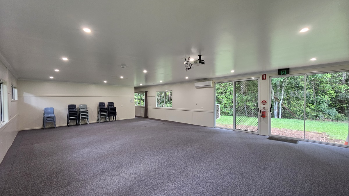A spacious open space with grey flooring and a white ceiling. Signs and a fire extinguisher are mounted on the middle wall, next to a glass door leading to the green field outside. A projector machine is mounted to the ceiling along with LED lights. Plastic chairs are piled against the middle wall. Trees are visible on the right. A spacious open space with grey flooring and a white ceiling. Signs and a fire extinguisher are mounted on the middle wall, next to a glass door leading to the green field outside. A projector machine is mounted to the ceiling along with LED lights. Plastic chairs are piled against the middle wall. Trees are visible on the right.