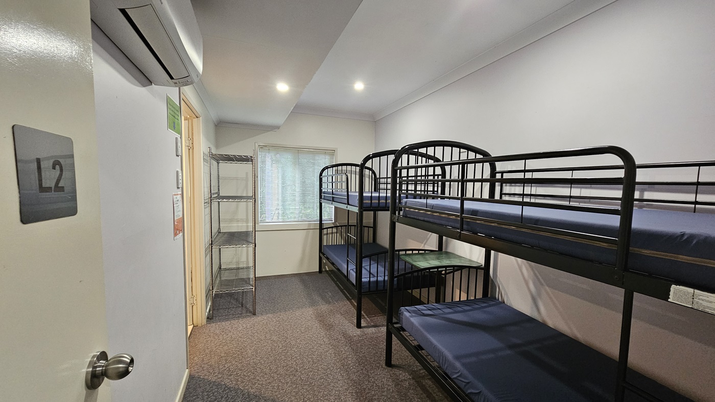 A room with metal bunk beds, each with mattresses, positioned against the wall. On the left is a door, adjacent to a 5-tier movable stainless display rack against the wall. In the middle wall is a glass window with blinds. The walls are white with poster reminders. An air conditioner is mounted to the upper left wall. The ceiling is white with fluorescent lights, and the floor is brown timber. A room with metal bunk beds, each with mattresses, positioned against the wall. On the left is a door, adjacent to a 5-tier movable stainless display rack against the wall. In the middle wall is a glass window with blinds. The walls are white with poster reminders. An air conditioner is mounted to the upper left wall. The ceiling is white with fluorescent lights, and the floor is brown timber.