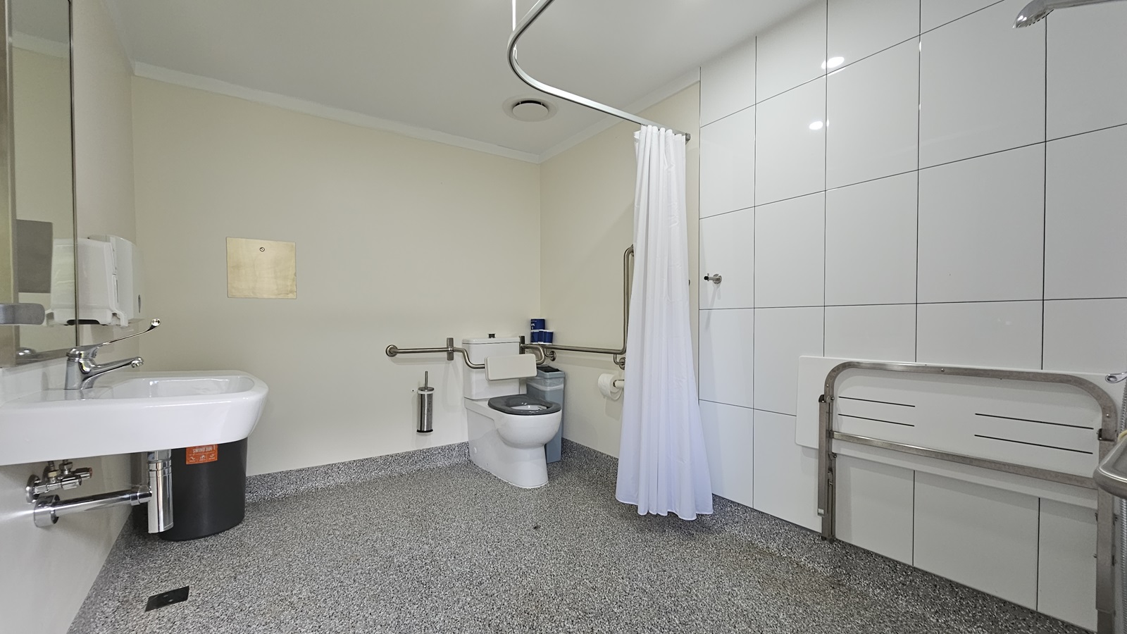 A spacious toilet with a shower. A hand basin, mirror, liquid soap, and tissue dispenser are mounted on the left wall, with a black rubbish bin adjacent. To the right is a toilet bowl equipped with hand grabs, with a toilet brush, rubbish bin, and tissue dispenser nearby. Adjacent is a shower and a foldable bench mounted to the right wall. A white shower curtain hangs from the ceiling. The walls and ceiling are all white, and the floor has grey tiles. A spacious toilet with a shower. A hand basin, mirror, liquid soap, and tissue dispenser are mounted on the left wall, with a black rubbish bin adjacent. To the right is a toilet bowl equipped with hand grabs, with a toilet brush, rubbish bin, and tissue dispenser nearby. Adjacent is a shower and a foldable bench mounted to the right wall. A white shower curtain hangs from the ceiling. The walls and ceiling are all white, and the floor has grey tiles.