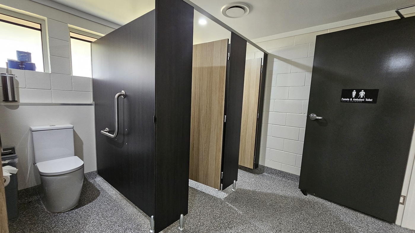 Three bathroom stalls, each with a black wooden door and timber partitions. To the right is a toilet bowl with handrails. To its left is a rubbish bin and tissue dispenser. Above is a toilet bowl brush and tissue rolls placed on a window ledge. The wall has white bricks, the ceiling is white with LED lights, and the floor has grey tiles. The inward-opening black door bears a sign indicating gender and accessibility use. Three bathroom stalls, each with a black wooden door and timber partitions. To the right is a toilet bowl with handrails. To its left is a rubbish bin and tissue dispenser. Above is a toilet bowl brush and tissue rolls placed on a window ledge. The wall has white bricks, the ceiling is white with LED lights, and the floor has grey tiles. The inward-opening black door bears a sign indicating gender and accessibility use.
