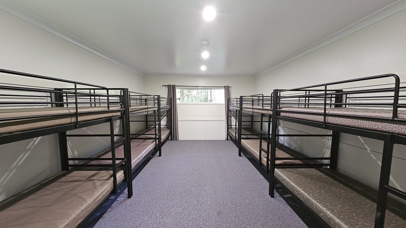 A room with metal bunk beds, each with mattresses, positioned against the walls. In the middle is a glass window with grey curtains pulled to the sides. The walls are white, the ceiling is white with fluorescent lights, and the floor is printed with grey tiles. A room with metal bunk beds, each with mattresses, positioned against the walls. In the middle is a glass window with grey curtains pulled to the sides. The walls are white, the ceiling is white with fluorescent lights, and the floor is printed with grey tiles.