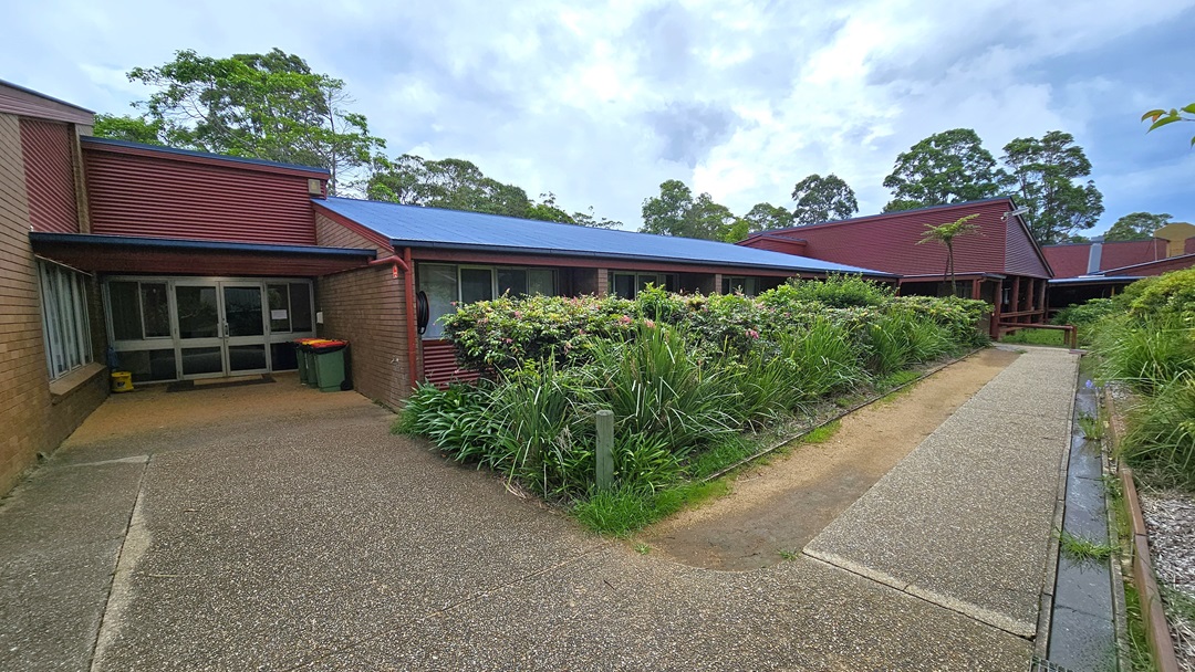 A shaded area with tiered seating extending to the right. Blue plastic chairs are positioned alongside with signage. To the left, another shade structure adjoins a low-rise building with a brown brick facade bearing the name "Merrin Lodge". The roofing structure features corrugated sheets and metal beams. Landscape ornamental plants occupy the front and sides of the building, with trees surrounding the area. A shaded area with tiered seating extending to the right. Blue plastic chairs are positioned alongside with signage. To the left, another shade structure adjoins a low-rise building with a brown brick facade bearing the name "Merrin Lodge". The roofing structure features corrugated sheets and metal beams. Landscape ornamental plants occupy the front and sides of the building, with trees surrounding the area.