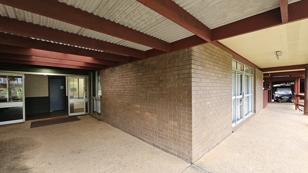 To the left, an outward-opening glass door equipped with matting. The walls have brown brick on the outside and white on the inside. The ceiling has exposed metal beams with a corrugated roof. On the right, a small vehicle is parked. To the left, an outward-opening glass door equipped with matting. The walls have brown brick on the outside and white on the inside. The ceiling has exposed metal beams with a corrugated roof. On the right, a small vehicle is parked.