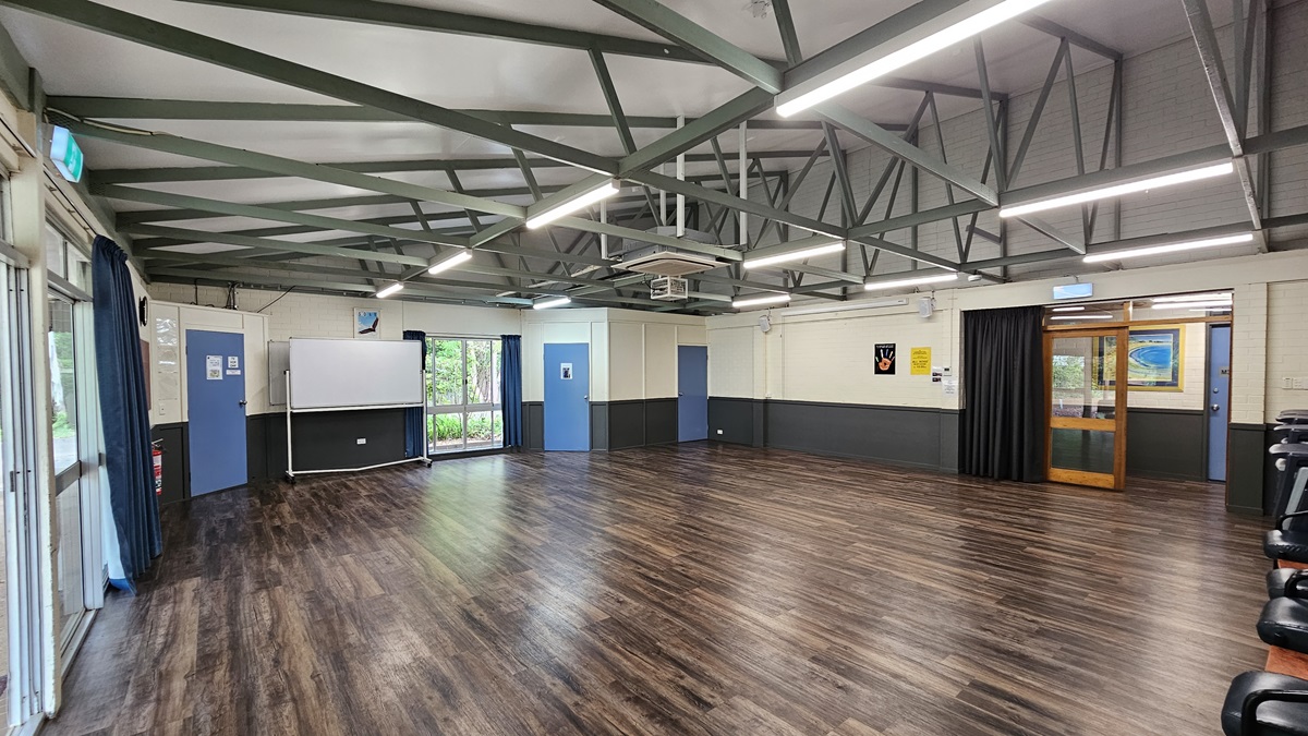 A spacious open area with a brown timber floor. The ceiling features white with exposed metal beams, fluorescent lights, air conditioning, and a projector machine. Glass windows have blue curtains rolled to the sides, next to a movable whiteboard. Blue doors and walls are equipped with posters. To the right is a glass door leading to corridors. Black chairs are arranged against the right wall. A spacious open area with a brown timber floor. The ceiling features white with exposed metal beams, fluorescent lights, air conditioning, and a projector machine. Glass windows have blue curtains rolled to the sides, next to a movable whiteboard. Blue doors and walls are equipped with posters. To the right is a glass door leading to corridors. Black chairs are arranged against the right wall.