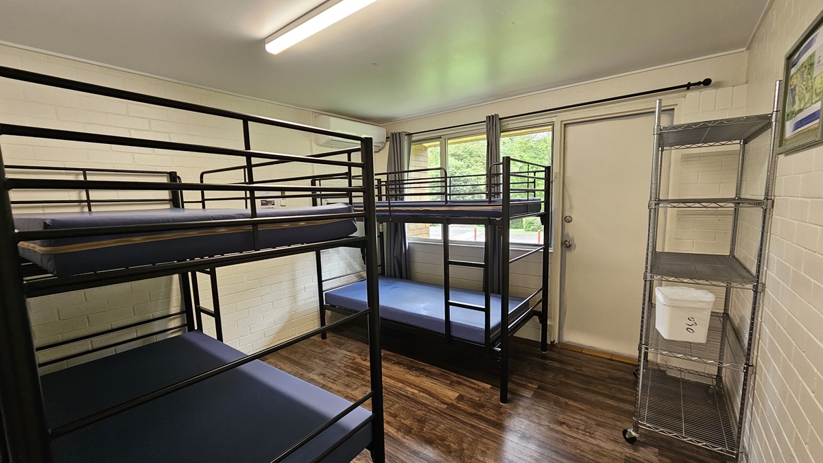 A room equipped with metal bunk beds, each with mattresses, positioned against the wall. On the right is a door beside a glass window with grey curtains rolled to the sides. Adjacent to it is a 5-tier movable stainless display rack against the wall. The walls are white with a picture frame decoration. The ceiling is white with fluorescent lights, and the floor is brown timber. A room equipped with metal bunk beds, each with mattresses, positioned against the wall. On the right is a door beside a glass window with grey curtains rolled to the sides. Adjacent to it is a 5-tier movable stainless display rack against the wall. The walls are white with a picture frame decoration. The ceiling is white with fluorescent lights, and the floor is brown timber.