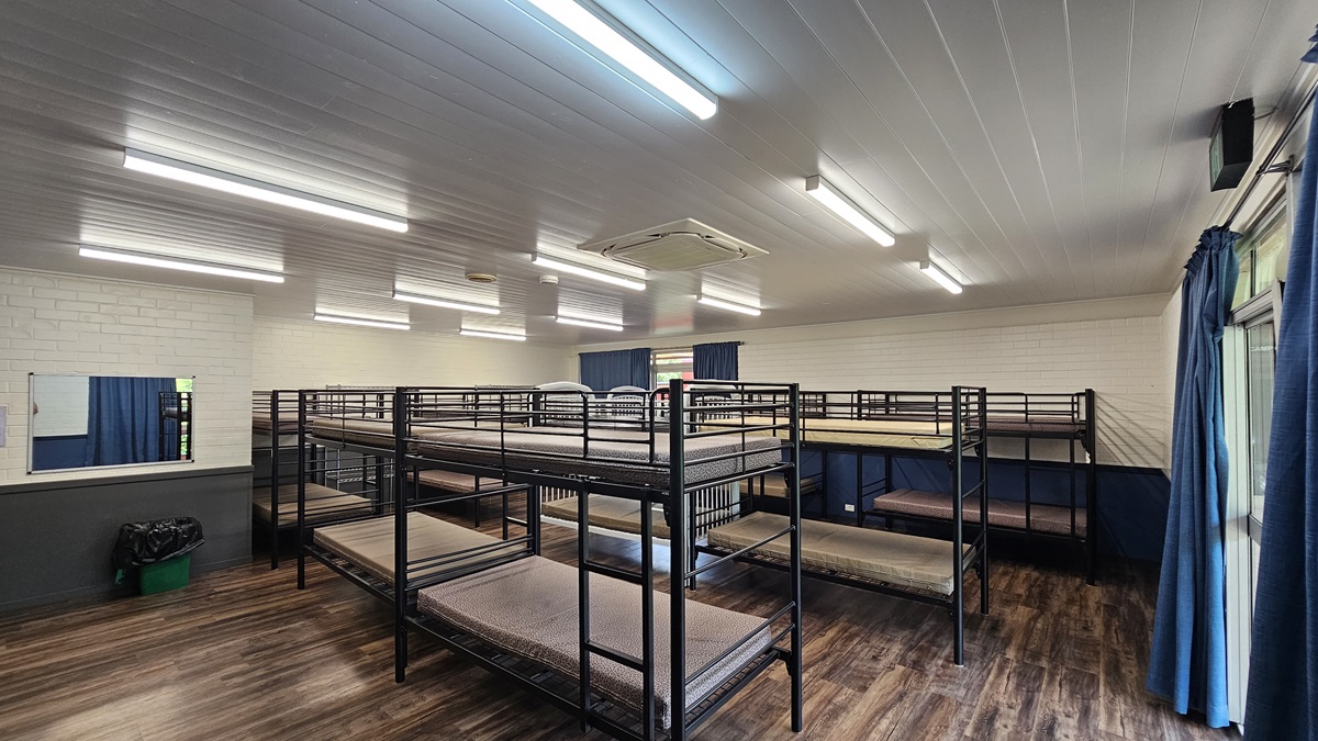 A room equipped with metal bunk beds, each with mattresses. On the right is a door beside a glass window with blue curtains rolled to the sides. The walls are white bricks with a mirror on the left and a rubbish bin below. The ceiling is white with fluorescent lights and air conditioning, and the floor is brown timber. A room equipped with metal bunk beds, each with mattresses. On the right is a door beside a glass window with blue curtains rolled to the sides. The walls are white bricks with a mirror on the left and a rubbish bin below. The ceiling is white with fluorescent lights and air conditioning, and the floor is brown timber.