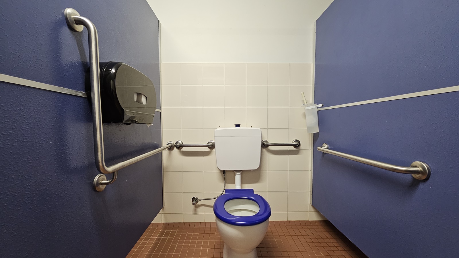 In the middle, a toilet bowl equipped with hand grabs. A tissue dispenser is mounted on the left wall. Blue walls on both sides, with white tiles on the back wall. A toilet brush is affixed to the upper right wall. The floor is tiled brown, and the ceiling is all white. In the middle, a toilet bowl equipped with hand grabs. A tissue dispenser is mounted on the left wall. Blue walls on both sides, with white tiles on the back wall. A toilet brush is affixed to the upper right wall. The floor is tiled brown, and the ceiling is all white.
