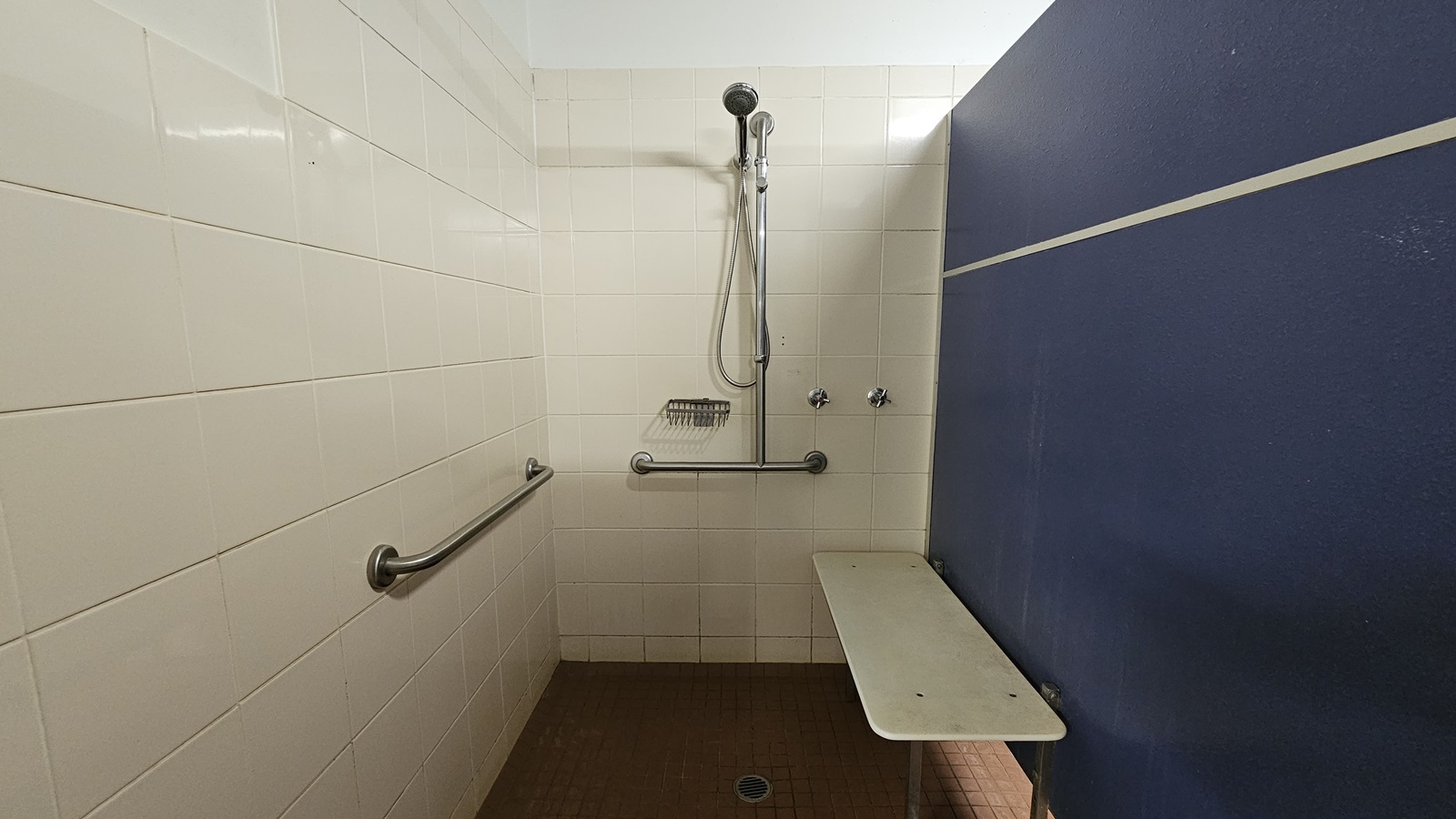 A shower with hand rails and a soap holder mounted on the middle wall. White tiles cover the left and middle walls, with a blue partition wall on the right. Brown tiles adorn the floor. The ceiling is white, and a foldable bench chair is mounted on the right wall. A shower with hand rails and a soap holder mounted on the middle wall. White tiles cover the left and middle walls, with a blue partition wall on the right. Brown tiles adorn the floor. The ceiling is white, and a foldable bench chair is mounted on the right wall.