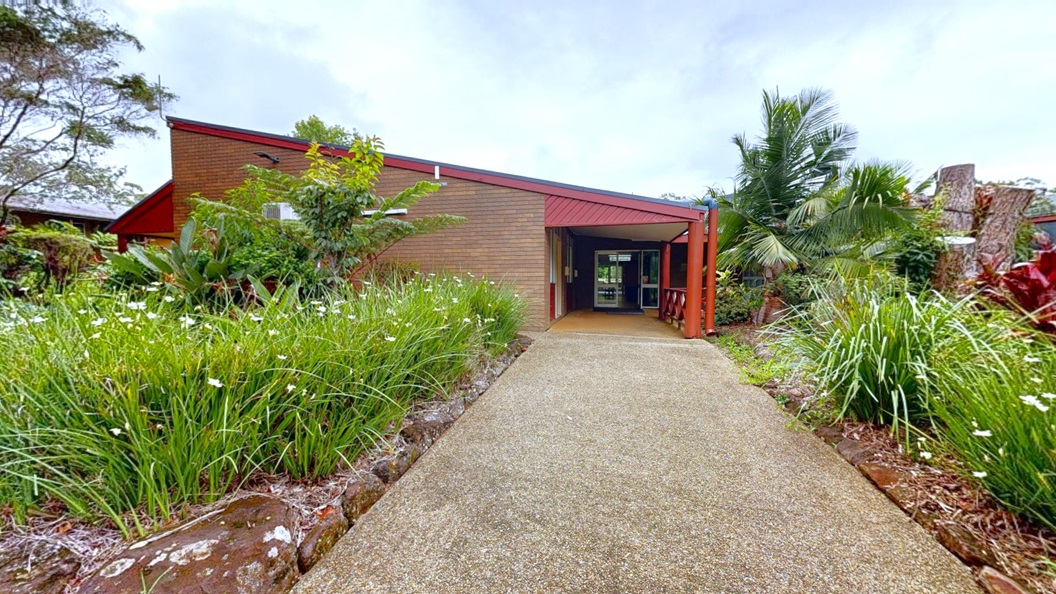 A concrete path leads towards a low-rise building with pitched roofing and a brown brick facade. The path is flanked on both sides by a landscape featuring flowering plants, large rocks, and various trees. A concrete path leads towards a low-rise building with pitched roofing and a brown brick facade. The path is flanked on both sides by a landscape featuring flowering plants, large rocks, and various trees.