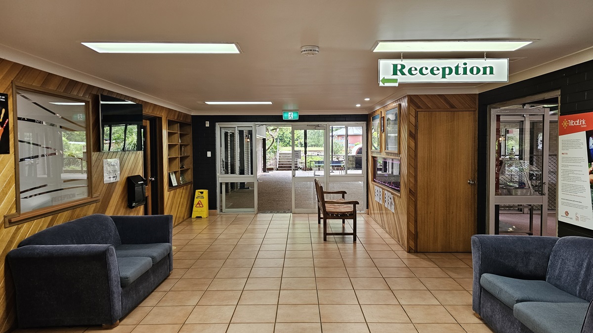 The interior has a low white ceiling with wooden accent walls. Grey sofas are placed on both sides. To the left is a door leading to the reception, where a hand sanitiser and a flatscreen TV are mounted on the wall. On the right, an inward-opening glass door is present. A wooden display cabinet, paintings, and an aquarium is positioned on the right with two chairs in front of it. Various signs are affixed to the wall and suspended from the ceiling. The interior has a low white ceiling with wooden accent walls. Grey sofas are placed on both sides. To the left is a door leading to the reception, where a hand sanitiser and a flatscreen TV are mounted on the wall. On the right, an inward-opening glass door is present. A wooden display cabinet, paintings, and an aquarium is positioned on the right with two chairs in front of it. Various signs are affixed to the wall and suspended from the ceiling.