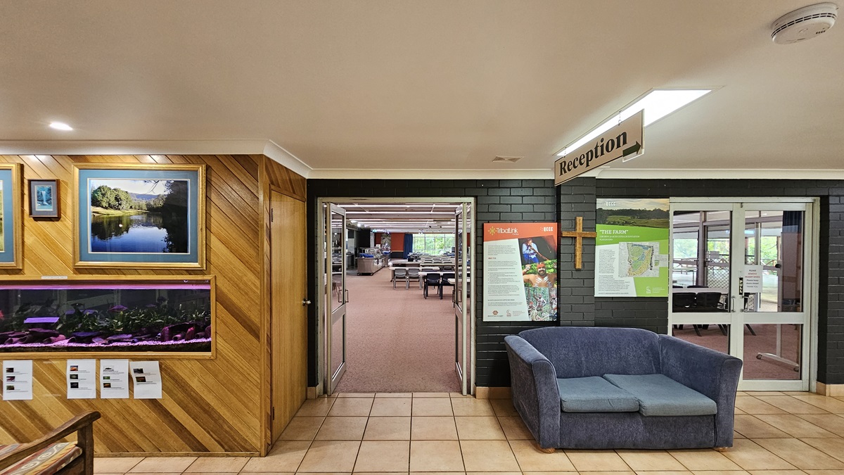On the left, there's a wooden display cabinet showcasing paintings and an aquarium, with two chairs positioned in front. To its right, an inward-opening glass door leads to the dining area, furnished with plastic chairs and tables. The flooring changes from cream outside to printed brown inside. Various signs are displayed on the walls and suspended from the ceiling. On the left, there's a wooden display cabinet showcasing paintings and an aquarium, with two chairs positioned in front. To its right, an inward-opening glass door leads to the dining area, furnished with plastic chairs and tables. The flooring changes from cream outside to printed brown inside. Various signs are displayed on the walls and suspended from the ceiling.