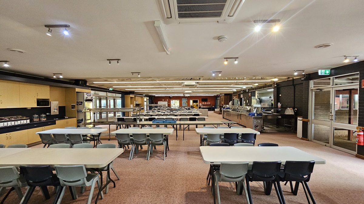 In the middle of the space, rows of tables and chairs are arranged. To the left, there is a cupboard filled with utensils. To the right is a section designated for food display. The low ceiling is white, equipped with LED and fluorescent lights, as well as air conditioning. The floor is adorned with printed brown tiles. On the right, a manual glass door serves as the exit, with a fire extinguisher and an exit sign mounted on the nearby wall. In the middle of the space, rows of tables and chairs are arranged. To the left, there is a cupboard filled with utensils. To the right is a section designated for food display. The low ceiling is white, equipped with LED and fluorescent lights, as well as air conditioning. The floor is adorned with printed brown tiles. On the right, a manual glass door serves as the exit, with a fire extinguisher and an exit sign mounted on the nearby wall.