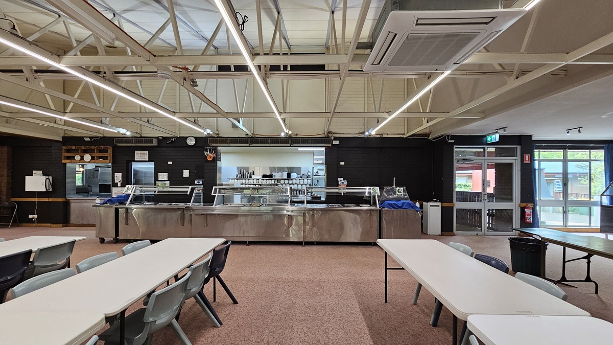 White plastic tables and chairs are neatly arranged in rows. A section for displaying food and utensils is visible in the background. The ceiling has exposed metal beams with fluorescent lights and air conditioning vents. The floor is covered with printed brown tiles. On the right, there's a manual glass door serving as an exit, with a fire extinguisher and an exit sign mounted on the nearby wall. White plastic tables and chairs are neatly arranged in rows. A section for displaying food and utensils is visible in the background. The ceiling has exposed metal beams with fluorescent lights and air conditioning vents. The floor is covered with printed brown tiles. On the right, there's a manual glass door serving as an exit, with a fire extinguisher and an exit sign mounted on the nearby wall.