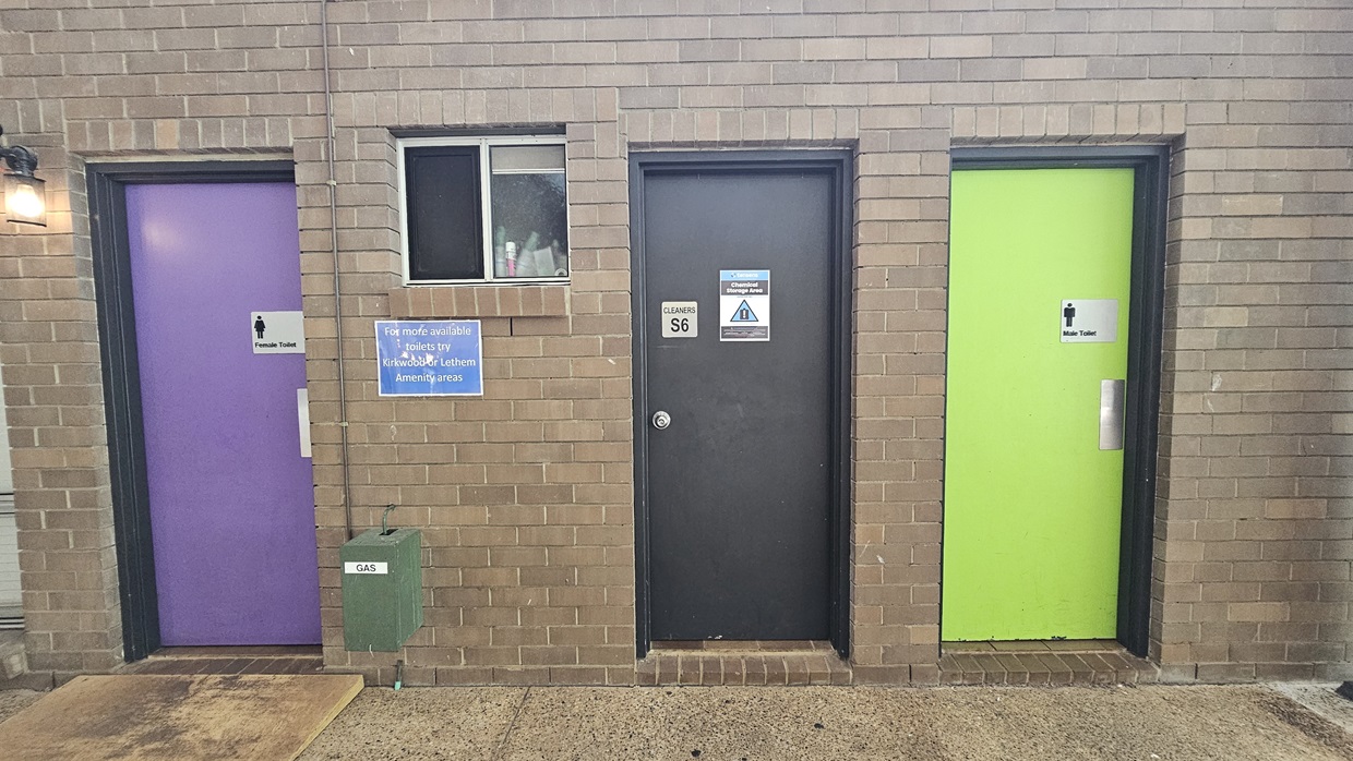 Three toilet doors of different colors: purple for Females, black for cleaning supplies access, and green for Males. Adjacent to the purple door is a window displaying toiletries, with signage. The wall is tiled in light brown, while the floor is cement. Three toilet doors of different colors: purple for Females, black for cleaning supplies access, and green for Males. Adjacent to the purple door is a window displaying toiletries, with signage. The wall is tiled in light brown, while the floor is cement.