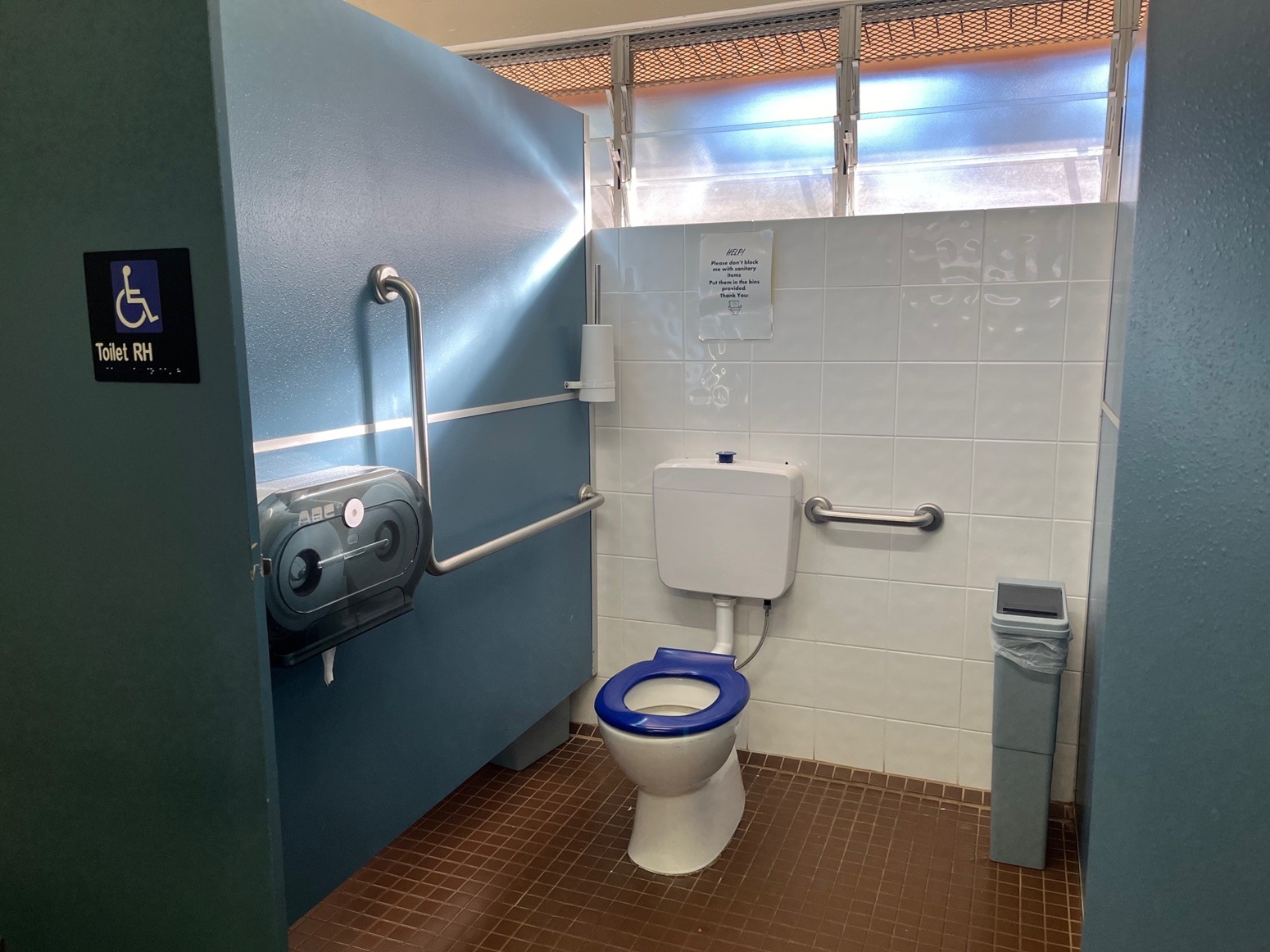 Inside the accessible toilet. A toilet bowl with handrails is in the left corner. A tissue roll dispenser is fixed to the blue wall. A toilet brush is mounted to the upper right blue wall. The back wall features white tiles and a glass window, while the floor is tiled brown. A reminder is affixed to the wall. Inside the accessible toilet. A toilet bowl with handrails is in the left corner. A tissue roll dispenser is fixed to the blue wall. A toilet brush is mounted to the upper right blue wall. The back wall features white tiles and a glass window, while the floor is tiled brown. A reminder is affixed to the wall.