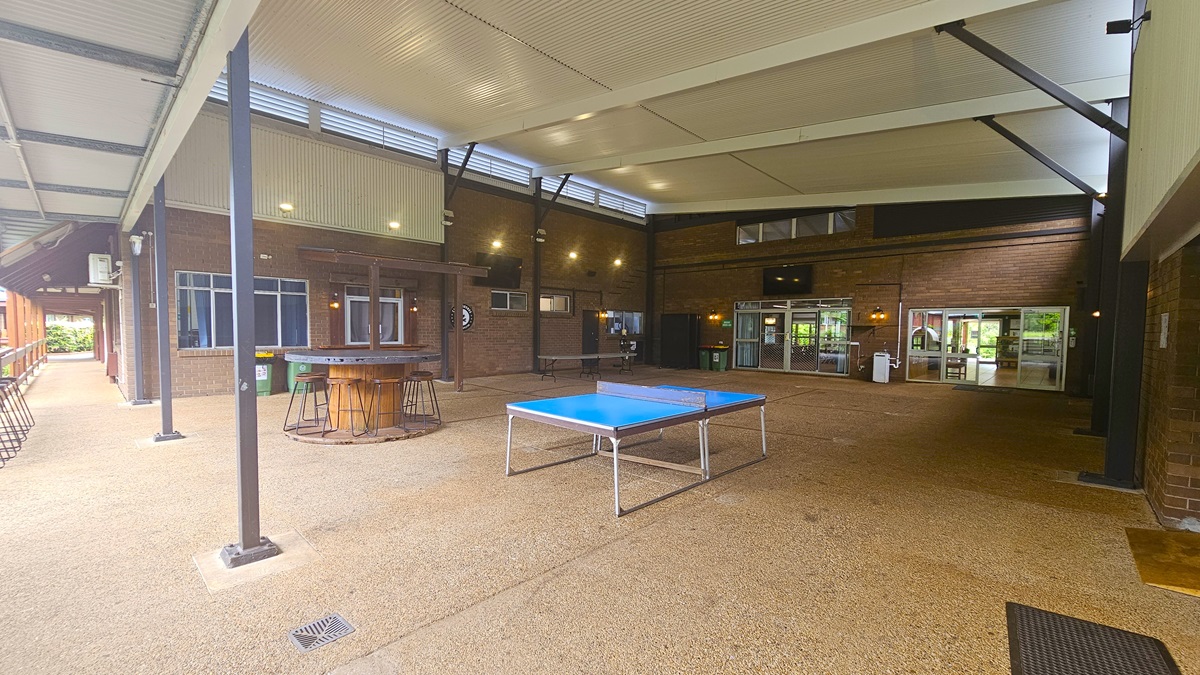 A spacious area with a round table and stools on the left, and a ping pong table in the middle-right. Brown brick walls and a cement floor with a sandy texture. White ceiling with exposed metal beams and corrugated roofing. A black mat is on the right. Flat-screen TVs are mounted on the upper middle and left walls, while white rubbish bins are below a window on the left. A spacious area with a round table and stools on the left, and a ping pong table in the middle-right. Brown brick walls and a cement floor with a sandy texture. White ceiling with exposed metal beams and corrugated roofing. A black mat is on the right. Flat-screen TVs are mounted on the upper middle and left walls, while white rubbish bins are below a window on the left.