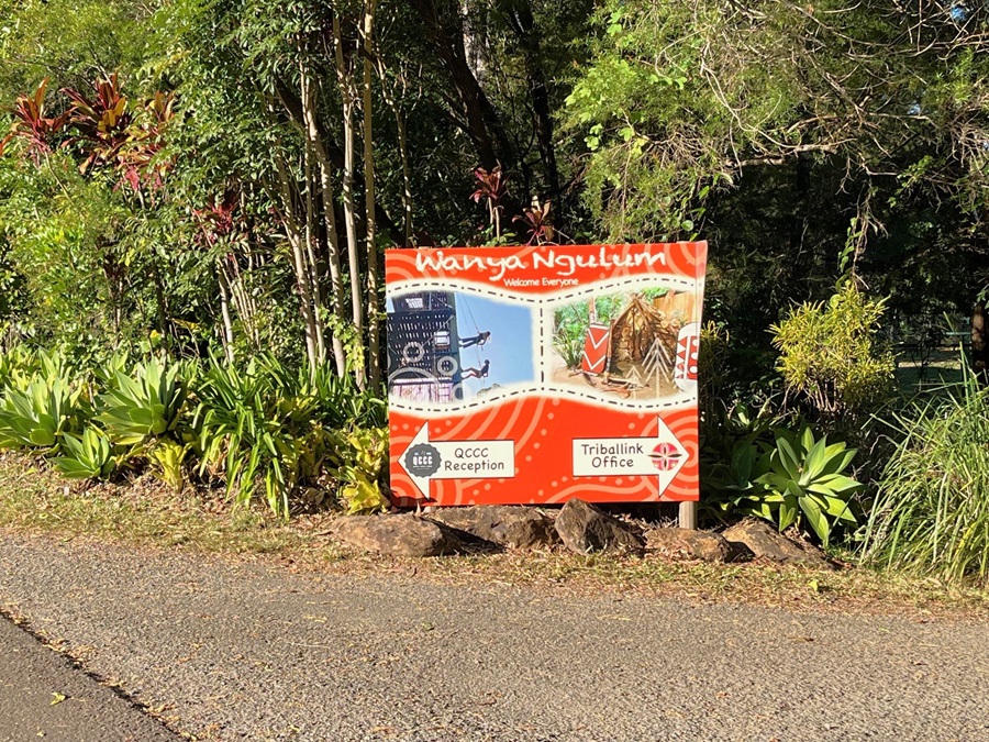 A concrete pathway slopes downward. On the right, a sign reading "TribalLink" is affixed to a wooden post alongside another signage. The path is bordered by plants, native grass, and trees. In the background, the roof of a facility is visible. A concrete pathway slopes downward. On the right, a sign reading "TribalLink" is affixed to a wooden post alongside another signage. The path is bordered by plants, native grass, and trees. In the background, the roof of a facility is visible.