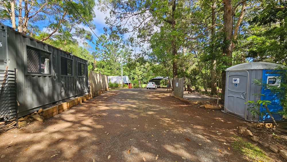 On the left stands a portable building with corrugated metal walls painted grey. Adjacent is a portable toilet, shaded by a tall tree. Further ahead is a dome-shaped structure and a white car. The area is surrounded by trees. On the left stands a portable building with corrugated metal walls painted grey. Adjacent is a portable toilet, shaded by a tall tree. Further ahead is a dome-shaped structure and a white car. The area is surrounded by trees.