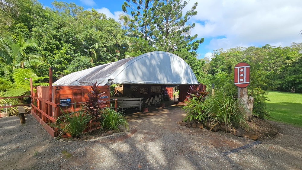 In the centre of a slightly elevated unpaved area stands a dome-shaped tent surrounded by a metal fence. Plants border the front. A signage resembling a traffic light is mounted on a tree trunk. Inside the tent are bench chairs and paintings on the wall. Trees and grass border the right side. In the centre of a slightly elevated unpaved area stands a dome-shaped tent surrounded by a metal fence. Plants border the front. A signage resembling a traffic light is mounted on a tree trunk. Inside the tent are bench chairs and paintings on the wall. Trees and grass border the right side.
