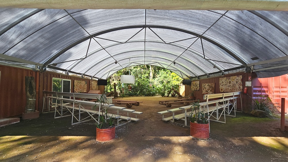 A dome-shaped tent with shade cloth roofing and unpaved ground. Inside, metal beams follow the contour. Tiered seating and improvised bench chairs are arranged on both sides with space in between. The red corrugated wall displays tribal paintings on the right. Indoor plants are placed in front and on the sides, with more visible behind. Flat-screen TVs and projection boards are also in place. A dome-shaped tent with shade cloth roofing and unpaved ground. Inside, metal beams follow the contour. Tiered seating and improvised bench chairs are arranged on both sides with space in between. The red corrugated wall displays tribal paintings on the right. Indoor plants are placed in front and on the sides, with more visible behind. Flat-screen TVs and projection boards are also in place.