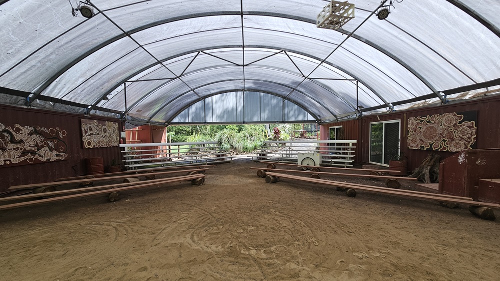 A dome-shaped tent with shade cloth roofing and unpaved ground. Inside, metal beams follow the contour. Tiered seating and improvised bench chairs are arranged on both sides with space in between. The red corrugated walls display tribal paintings. Indoor plants are placed in front and on the sides, with more visible behind. Flat-screen TVs and projection boards are also in place. A dome-shaped tent with shade cloth roofing and unpaved ground. Inside, metal beams follow the contour. Tiered seating and improvised bench chairs are arranged on both sides with space in between. The red corrugated walls display tribal paintings. Indoor plants are placed in front and on the sides, with more visible behind. Flat-screen TVs and projection boards are also in place.