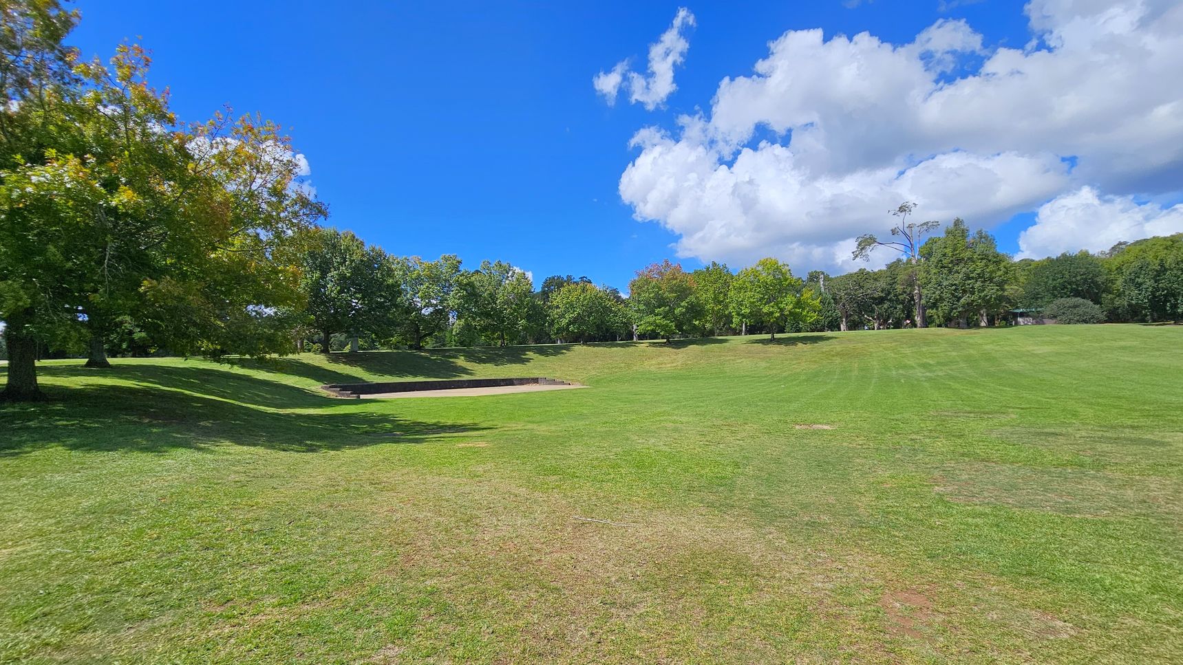 A grassy park landscape with a gentle slope. On the left, there are several large trees with green and yellow leaves, casting shadows on the ground. In the centre, a small, rectangular concrete structure is embedded in the grass. The right side features more open grassy space with a few scattered trees in the distance. The background is lined with a variety of trees, creating a natural border.
