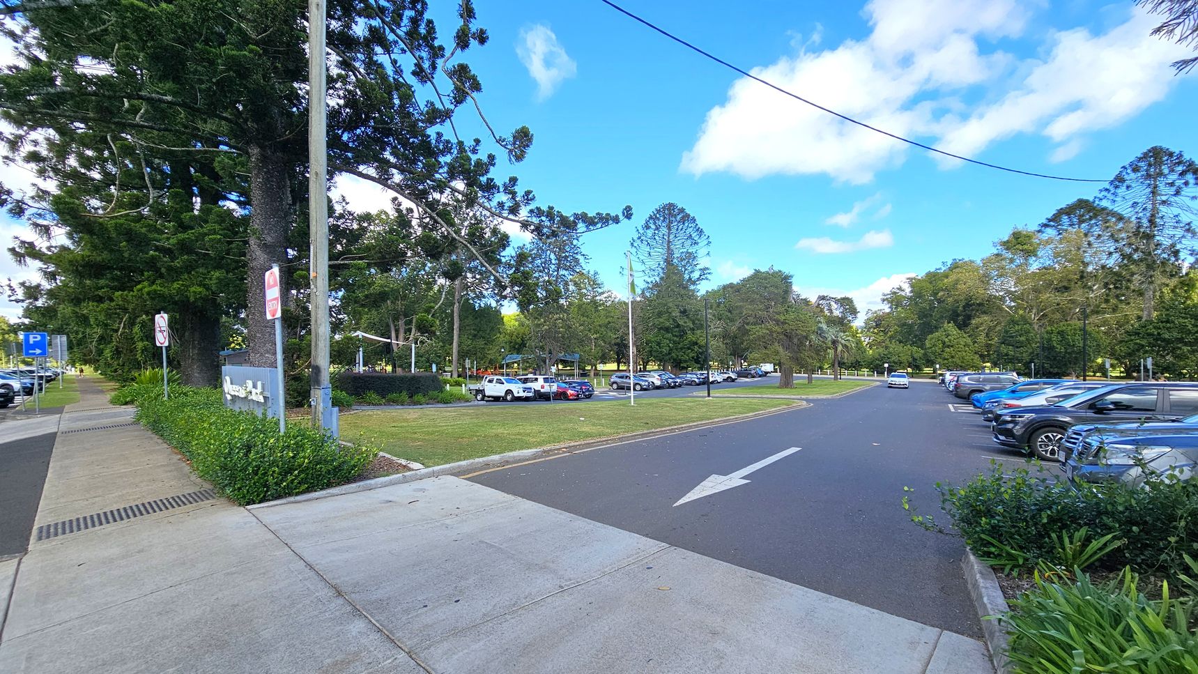 A paved footpath runs along the left side, bordered by a grassy area with large trees and a few signs. On the right, a car park is filled with vehicles, including a white truck and several blue cars. The road has a white arrow indicating direction. In the background, more trees and a park-like setting are visible. The sky is partly cloudy.