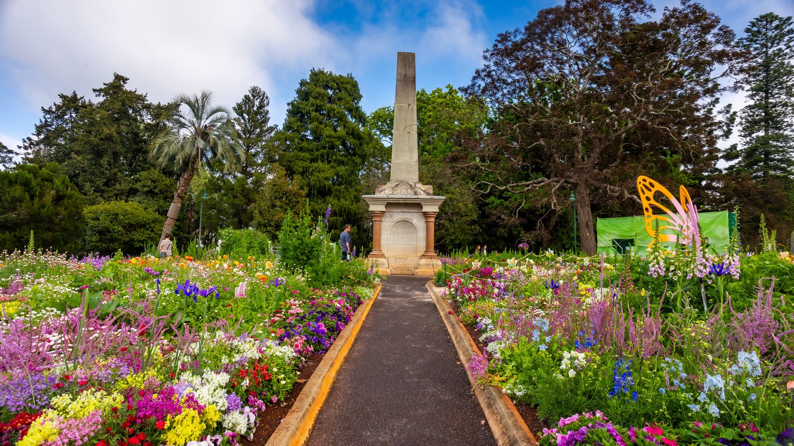 A vibrant garden with a central path leading to a tall stone obelisk. The path is bordered by colourful flowers in shades of purple, pink, yellow, and white. On the right, a large orange butterfly sculpture is visible. Tall trees, including a palm, form the background, creating a lush, green setting. The garden is well-maintained, with a mix of flowering plants and greenery.
