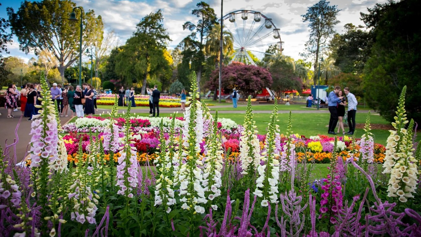 In the foreground, tall foxglove flowers in shades of purple and white stand prominently. Behind them, a variety of colourful flower beds are arranged in patterns. People are gathered on the left, some walking and others standing in groups. In the background, a Ferris wheel is visible among tall trees, adding a festive atmosphere to the park setting. The scene is lively with greenery and vibrant floral displays.