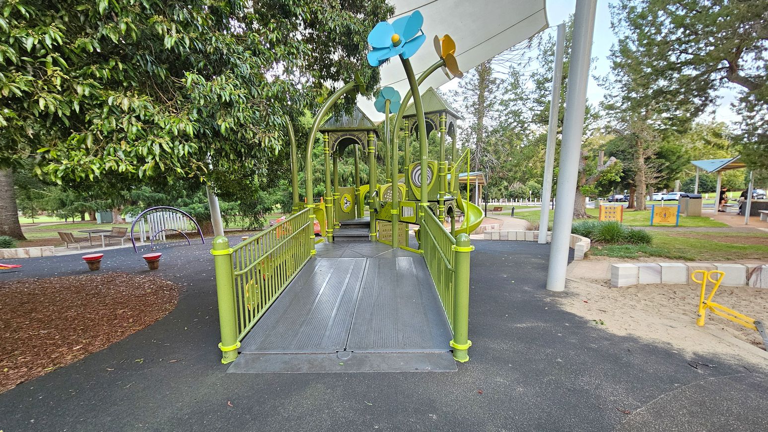 A playground with a green structure featuring a ramp leading to a play area with colourful flower decorations on top. On the left, there are trees and a curved climbing frame with benches nearby. The ground is covered with a mix of rubber and mulch. On the right, there's a sandy area with a yellow seesaw and a grassy section with picnic tables under a shelter. Trees and a car park are visible in the background.