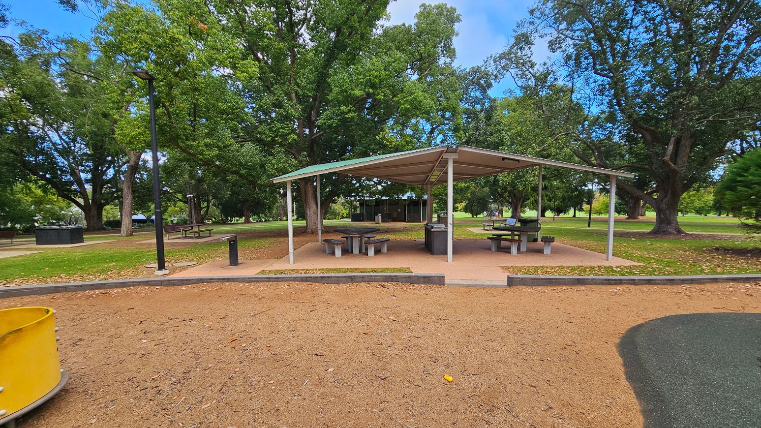 A park scene with a covered picnic area in the centre, featuring several tables and benches. The ground is a mix of grass and dirt, with a paved path leading to the shelter. A yellow play structure is partially visible on the left. Tall trees with dense green foliage surround the area, providing shade. A black lamp post stands near the shelter. In the background, more trees and open grassy spaces are visible.